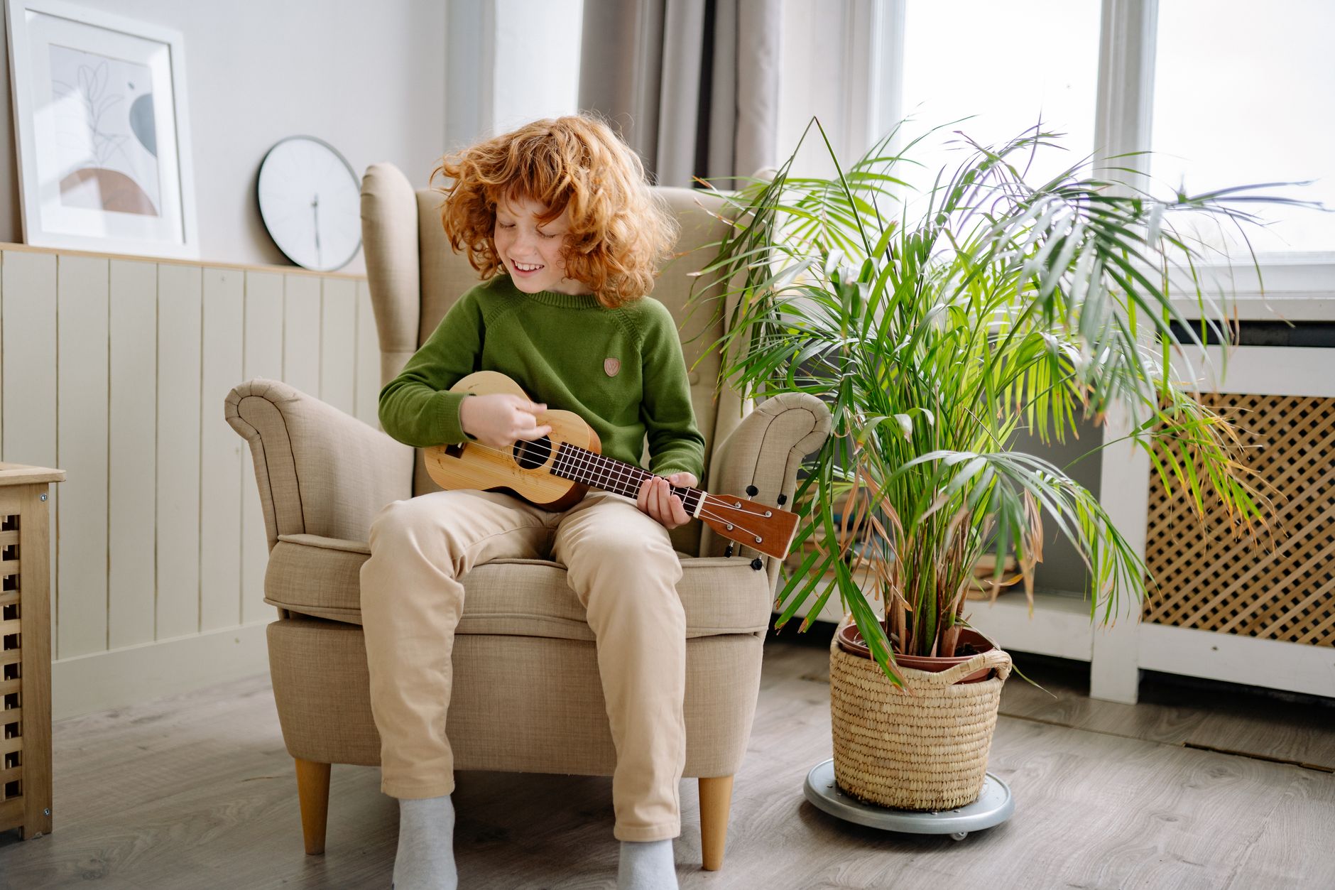 Red-haired child playing ukulele while sitting in a cozy armchair indoors with a houseplant nearby.
