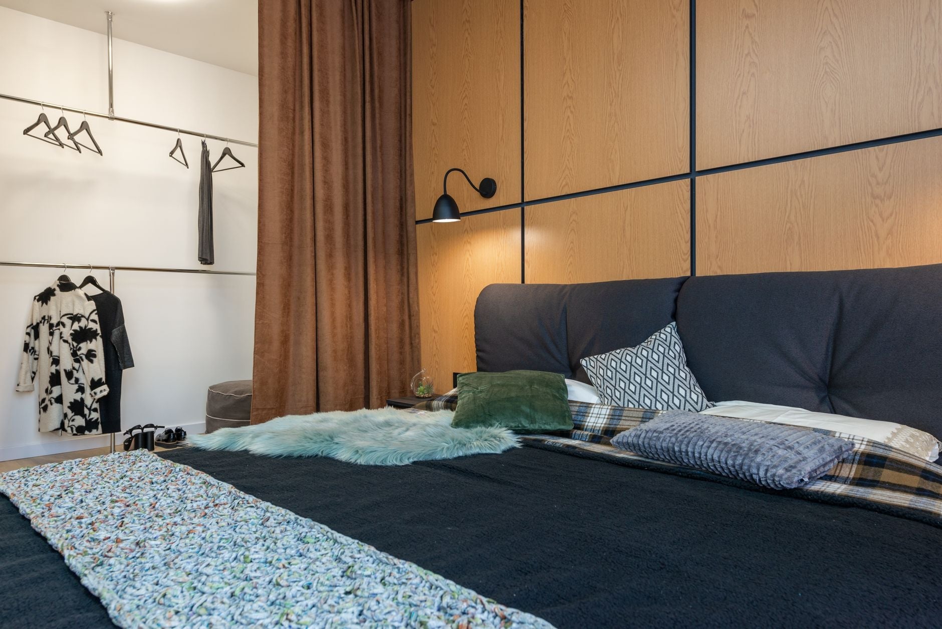 Comfortable bedroom with soft cushions on big bed placed near clothes hanging in wardrobe