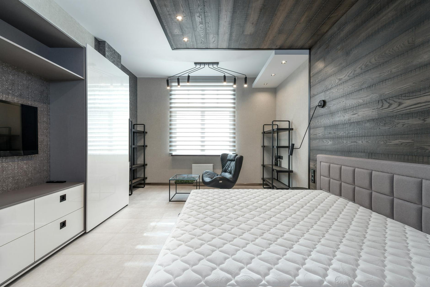 Bed with mattress placed against wall with modern TV and white cupboards in stylish bedroom with window and leather armchair
