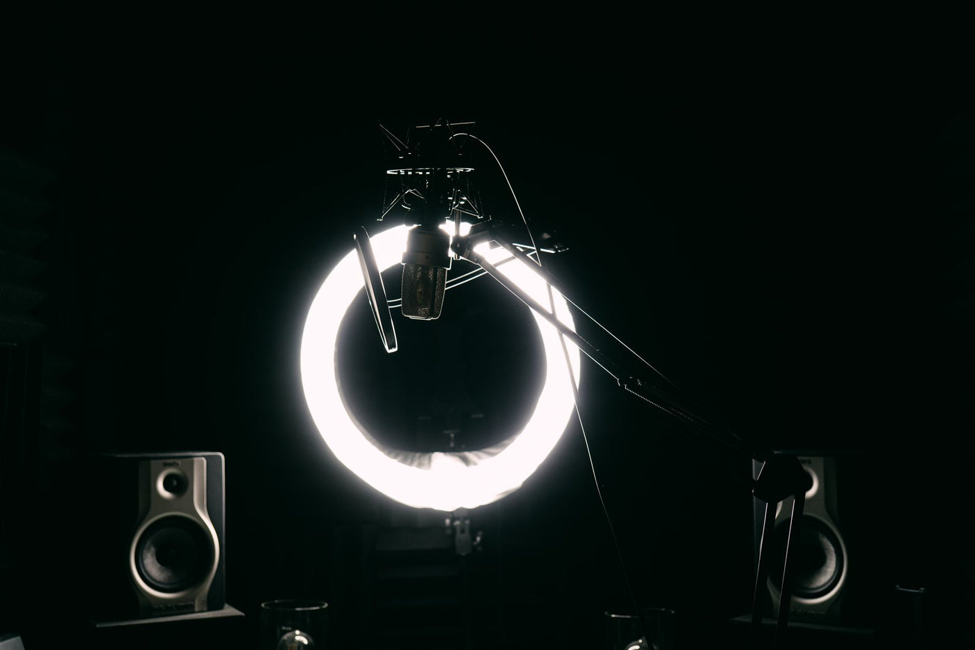 Dark studio room with condenser microphone, ring light, and speakers for recording purposes.