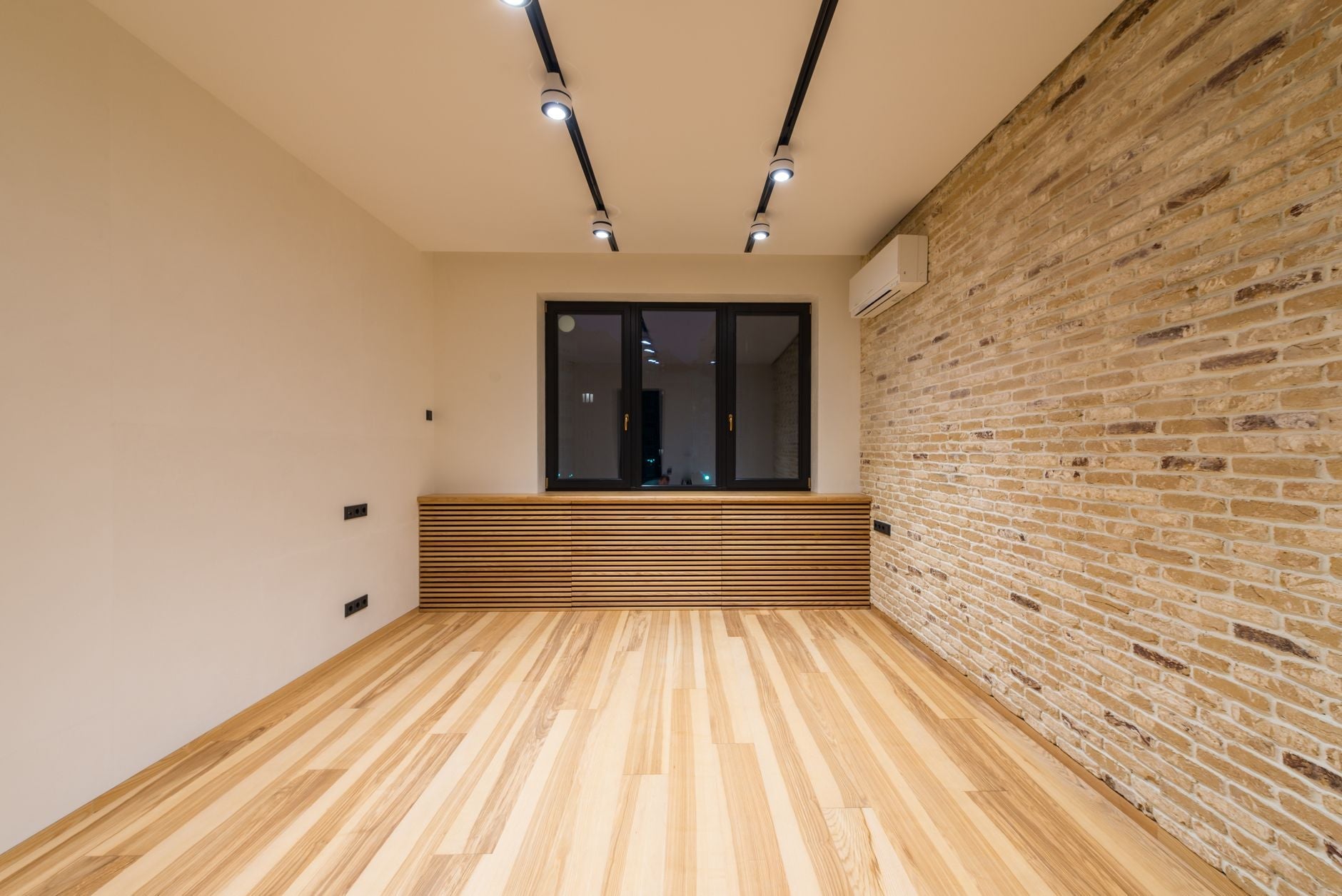 Empty modern room featuring wooden flooring, brick accent wall, and recessed lighting.