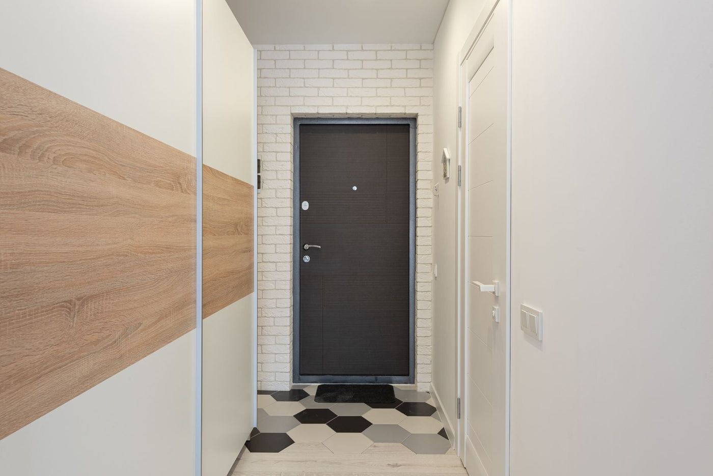 Entrance door between white brick wall against narrow passage with closet and rug with hexagonal ornament in house